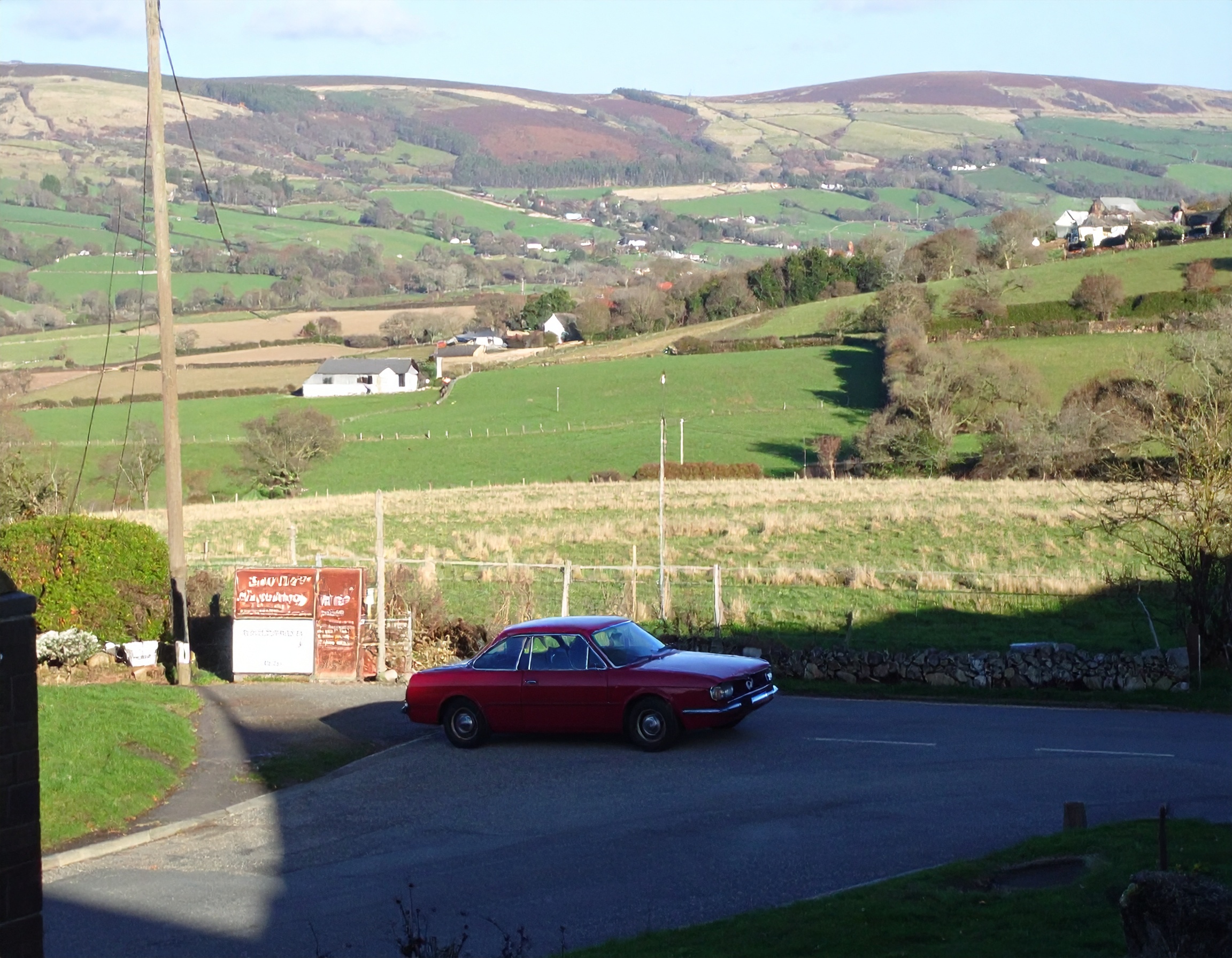 Classic red car in rural UK setting for "SellAVehicle.co.uk". Rolling green hills, fields, and farmhouses in the background.