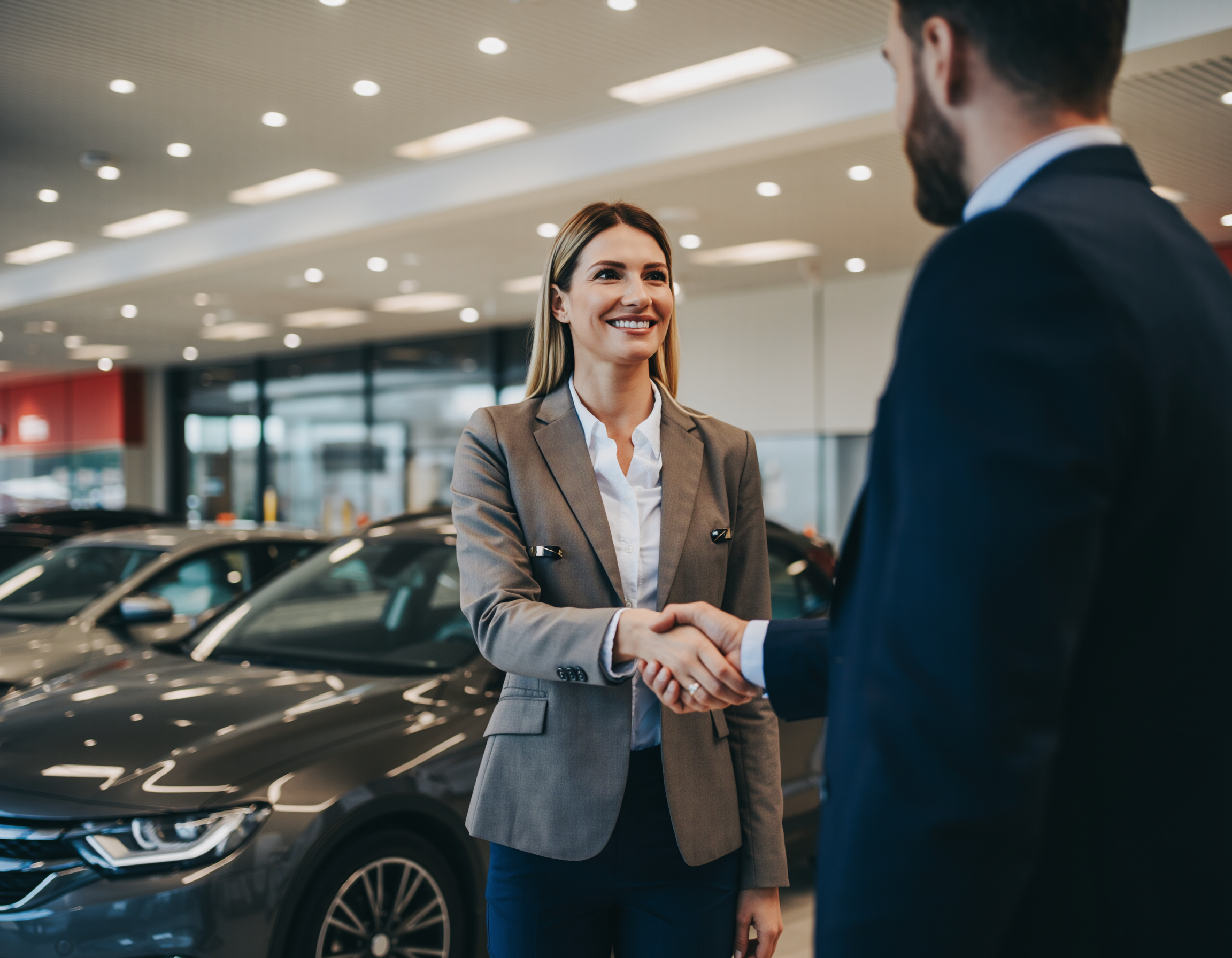 A smiling saleswoman shakes hands with a customer in a car dealership showroom.